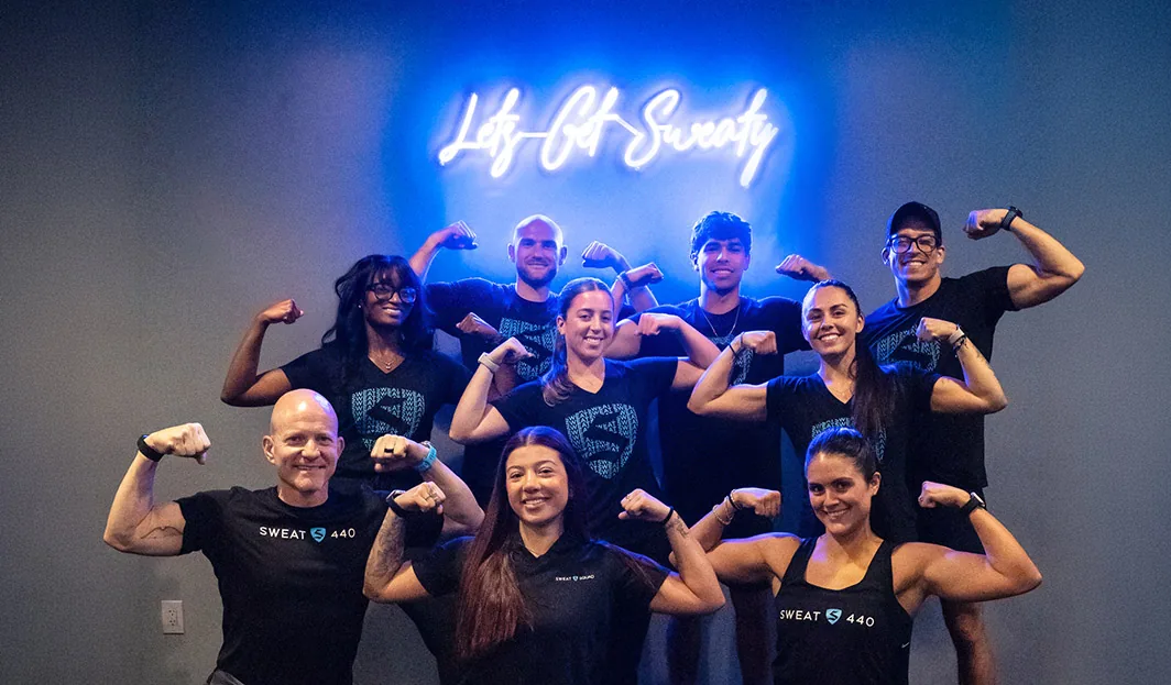 SWEAT440 coaches and members flexing together in front of the studio neon sign